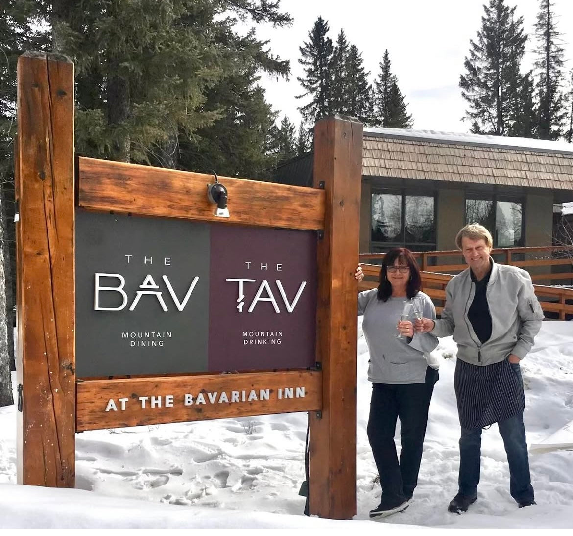 Two people standing next to a wooden sign for 'The Bav Tav' at The Bavarian Inn at Bragg Creek, Alberta, Canada