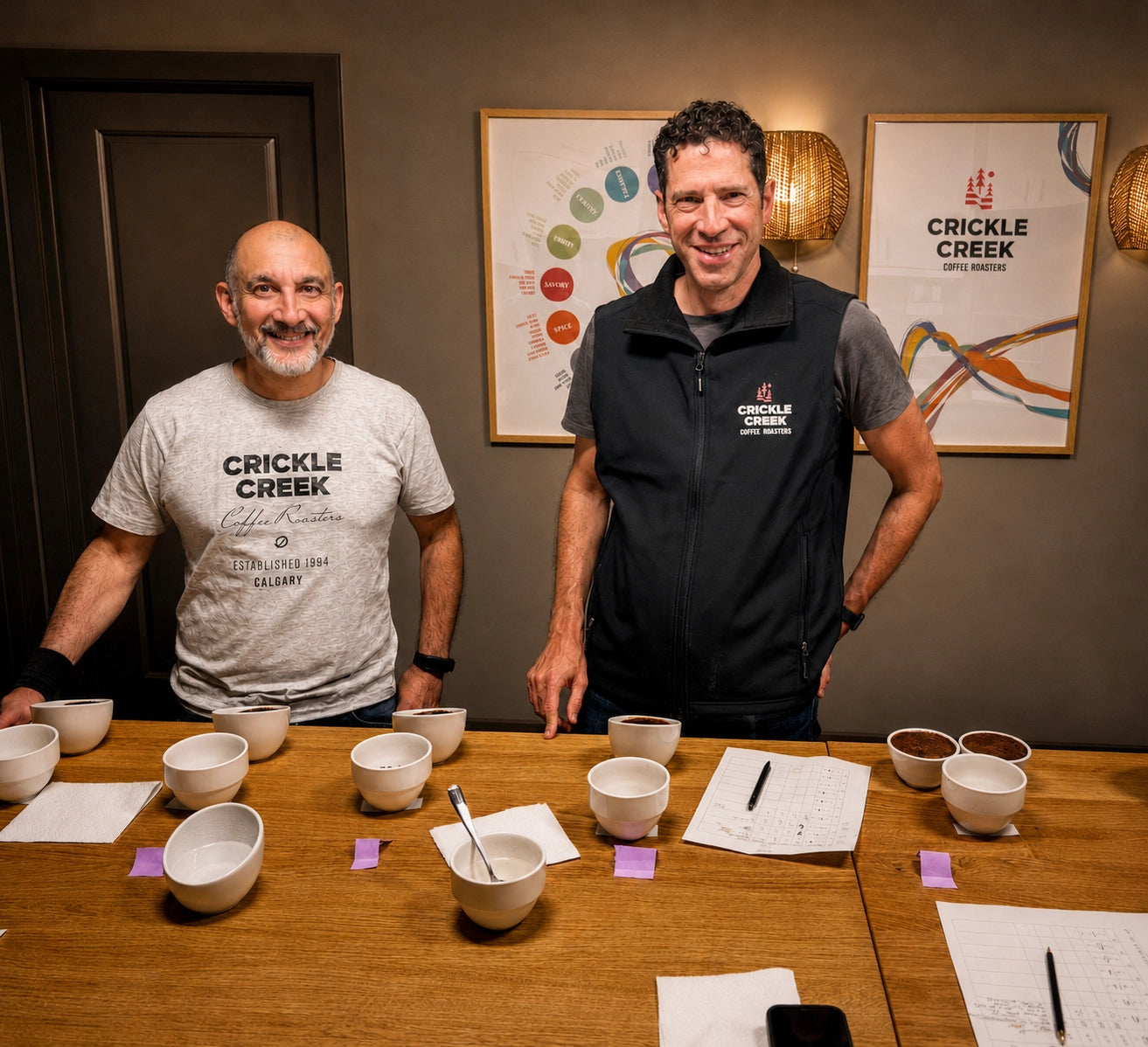 Two men standing behind a coffee cupping table with bowls and documents, with 'Crickle Creek' branding in the background.