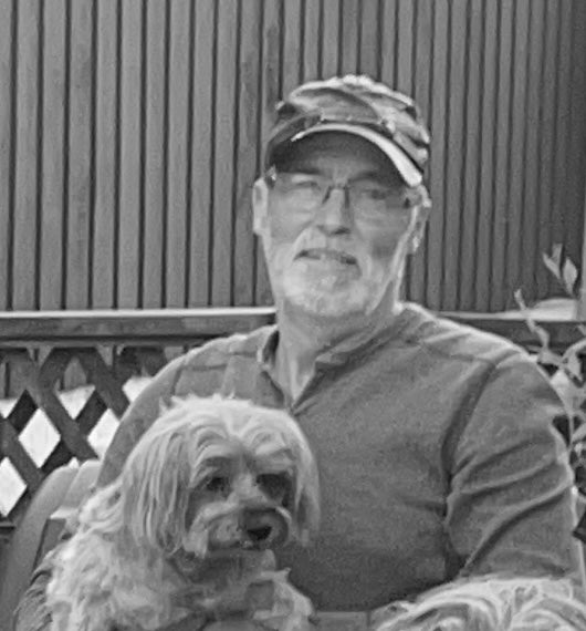 Man with a cap and glasses sitting next to a small dog outdoors.